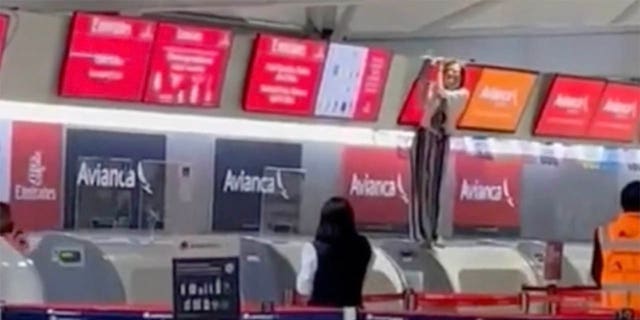 Otros pasajeros en el aeropuerto pueden ver a la persona fuera de control, de pie en el mostrador de facturación y sosteniendo una pantalla sobre ella, desde lejos.